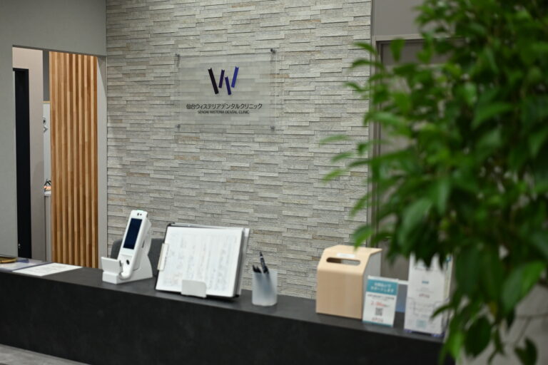Reception area of a dental clinic with a stone wall and a clear sign reading 'SENDAl WISTERIA DENTAL CLINIC' in Japanese and English, foreground shows desks and equipment, blurred green plant on right