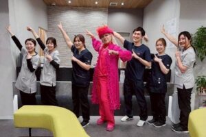 Group of healthcare workers in scrubs posing with a person in a bright pink costume in a playful dance pose in a clinic setting.
