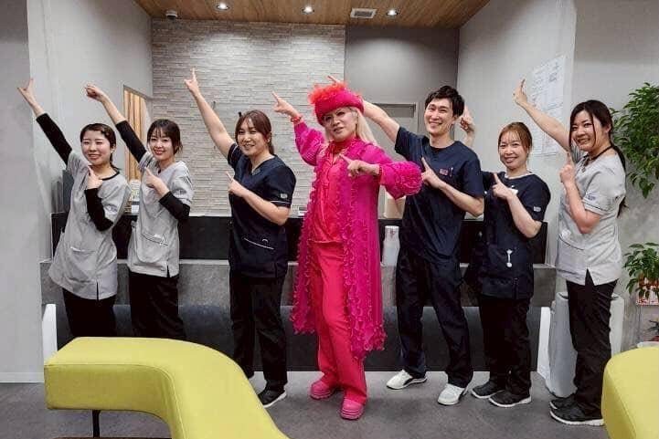 Group of healthcare workers in scrubs posing with a person in a bright pink costume in a playful dance pose in a clinic setting.
