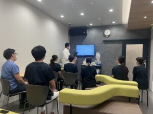People wearing masks seated in a room watching a presenter at a screen with a clock on the wall behind them.
