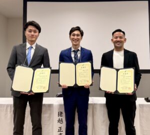 Three men in suits stand side by side, each holding an open certificate at an awards ceremony, smiling for the photo.