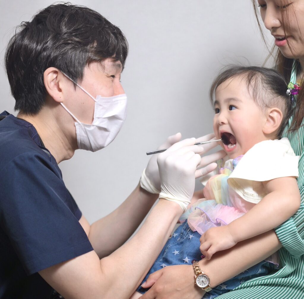 Dentist in a mask and gloves examining a toddler's open mouth with a dental tool, while a mother holds the child.