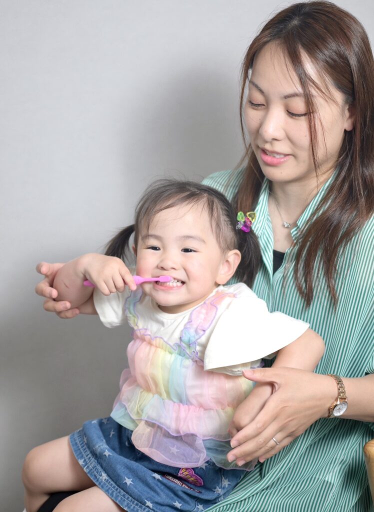 A smiling young child brushing teeth with a pink toothbrush, held by a woman in a green striped shirt.