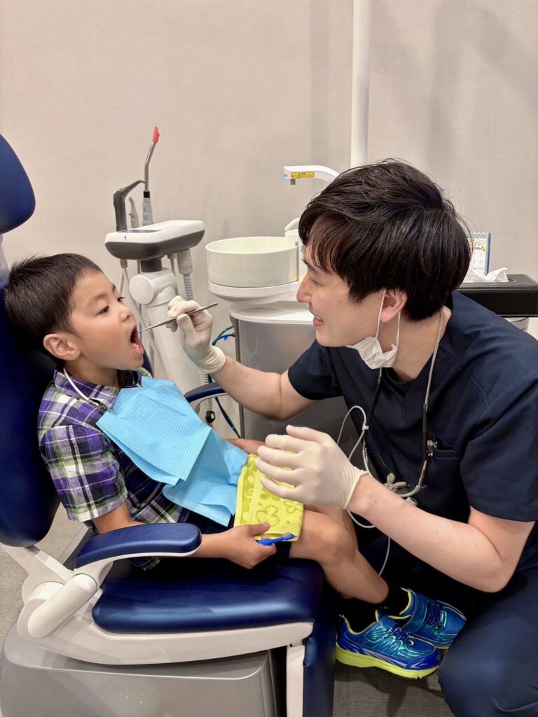 Dentist wearing gloves and mask examines a young boy's mouth in a dental chair, with a blue bib on the child and dental equipment nearby.