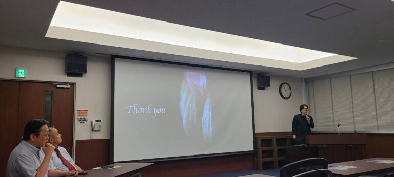 Speaker at a podium giving a presentation in a conference room, projection screen shows a slide with 'Thank you' text and a faded image behind it.