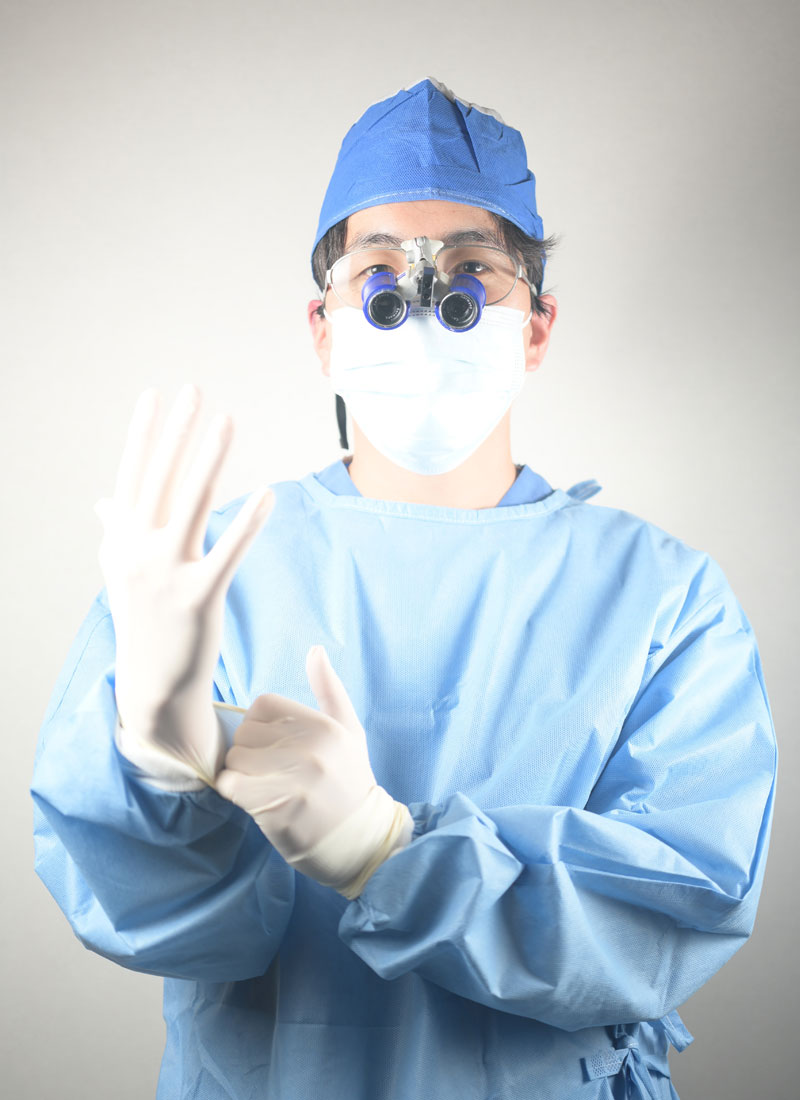 Surgeon in blue scrubs and a mask showing a stop gesture with gloved hands and loupes on to indicate caution or triage.