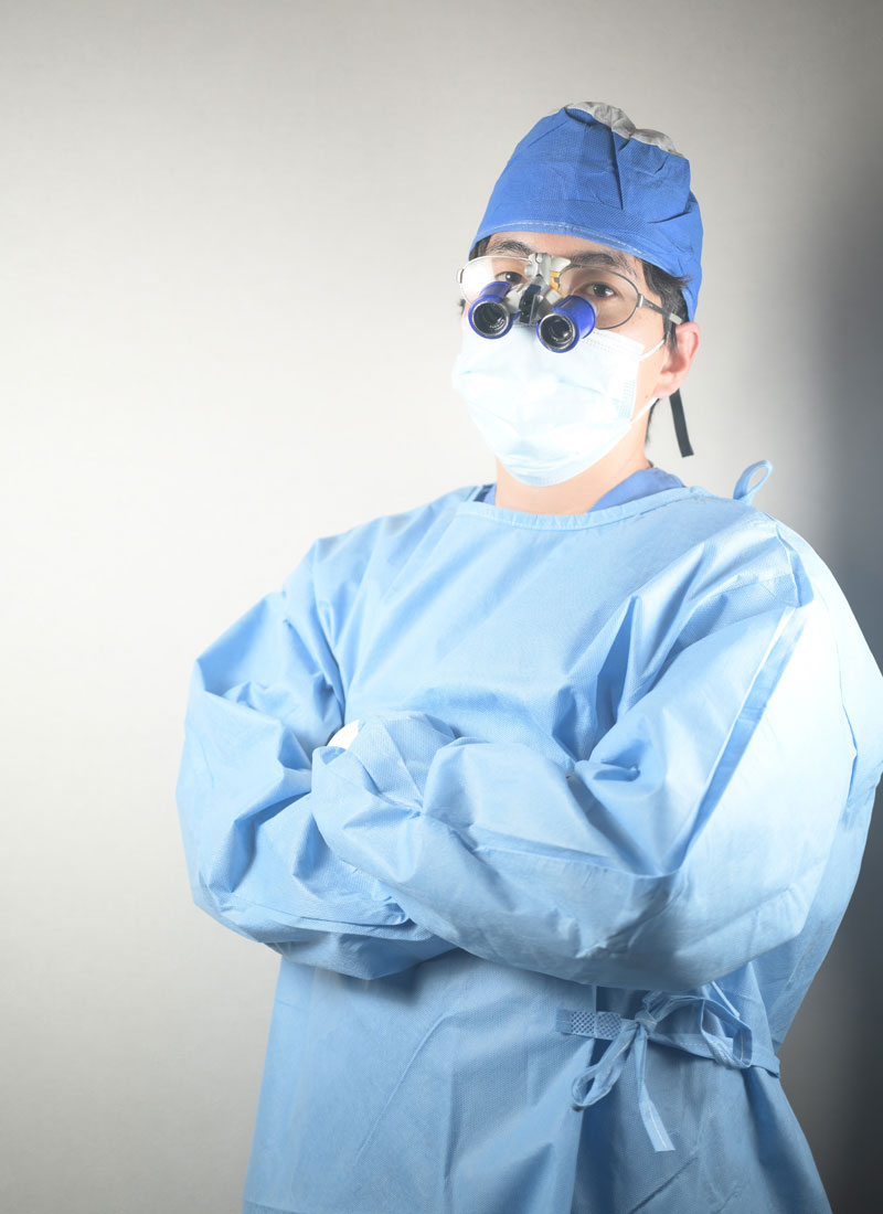 Surgeon in blue scrubs with mask and surgical cap, arms crossed, wearing magnifying loupes.