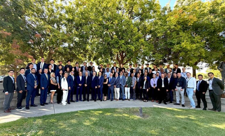 Large group of professionals in business attire posing together on outdoor steps under green trees for a group photo at an event.